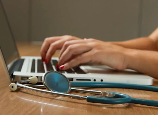 a physician typing on her laptop with her stethoscope on the side